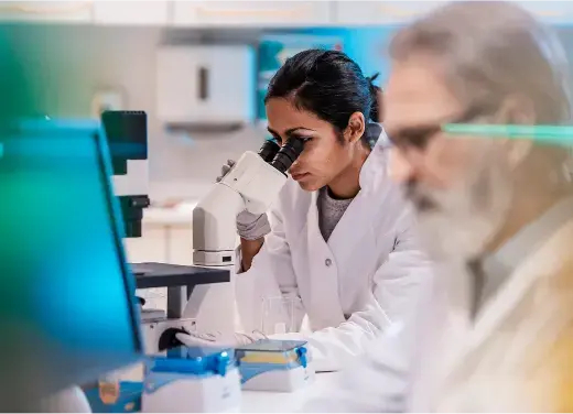Two scientists, one looking through a microscope, and one looking at a lab samples Two scientists, one looking through a microscope, and one looking at a lab samples